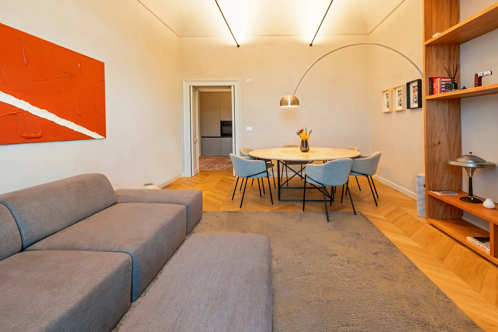 Divano grigio sotto un grande quadro arancione, con la zona pranzo sullo sfondo