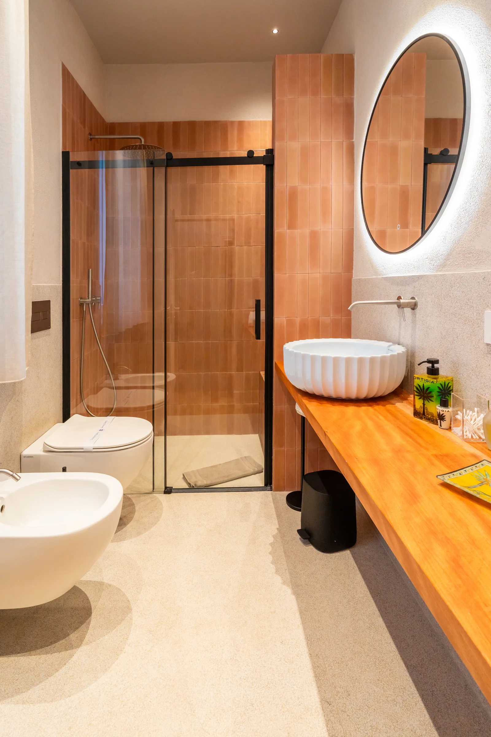 Bagno con doccia in piastrelle terracotta, specchio tondo retroilluminato e lavabo a coppa su piano in legno
